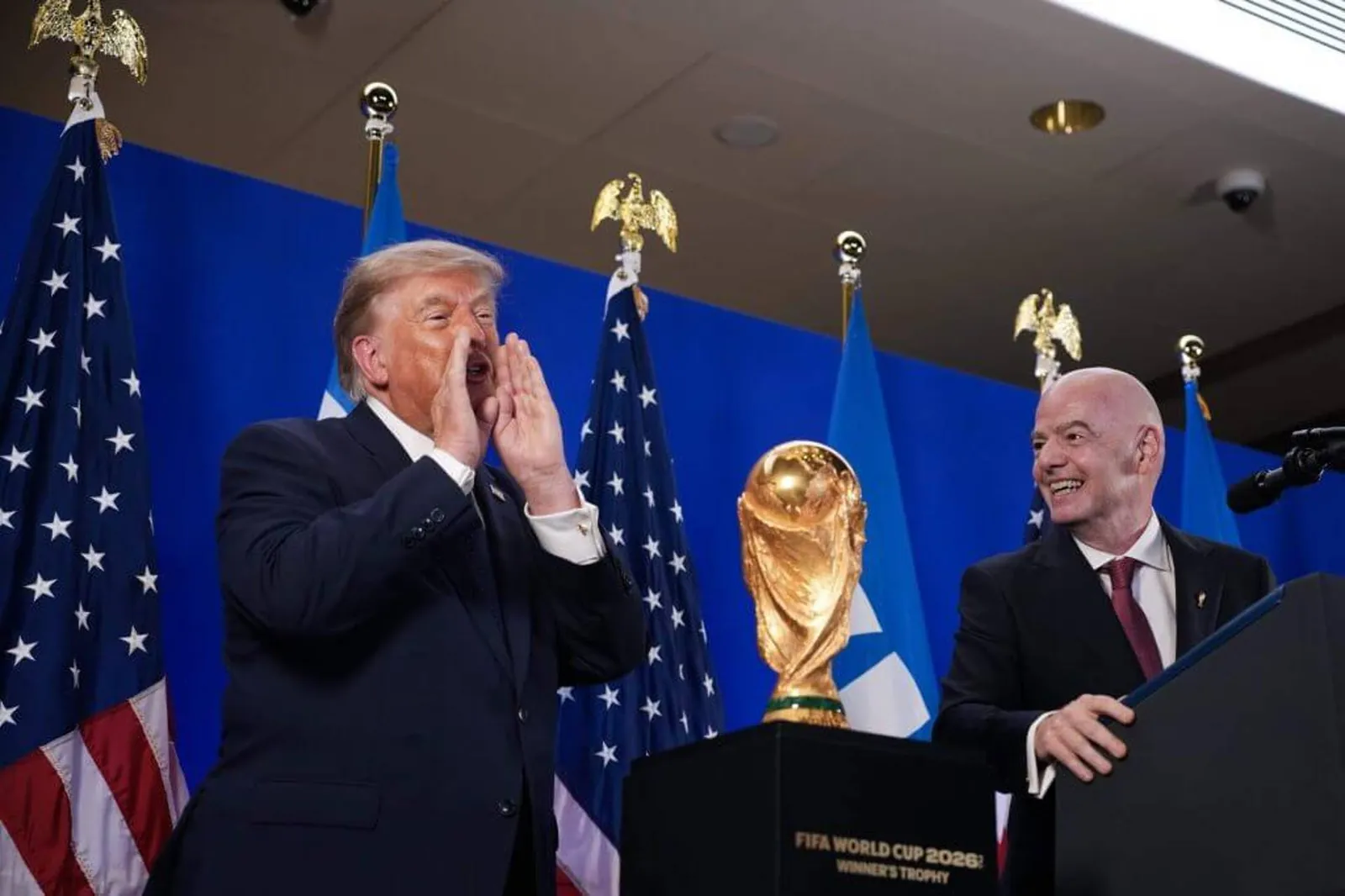 Trump with his hands to his mouth as if shouting. Infantino looks on, laughing.